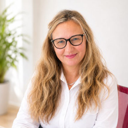Porträt einer lächelnden Frau mit Brille und weißem Hemd vor hellem Hintergrund